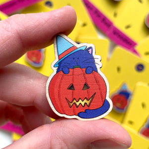 A wooden pin badge featuring a purple cat wearing a blue witches hat and standing in front of a lit halloween pumpkin is held between finger and thumb